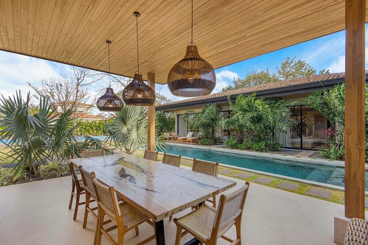 Een terras buiten met een houten tafel en stoelen, een zwembad en weelderige begroeiing.