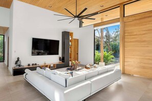Un salon moderne avec un grand canapé blanc, un téléviseur à écran plat, un ventilateur de plafond et une vue sur la verdure à l’extérieur.