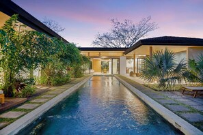 Une maison moderne dotée d’une piscine centrale, entourée d’une végétation luxuriante et sous un ciel dégagé.