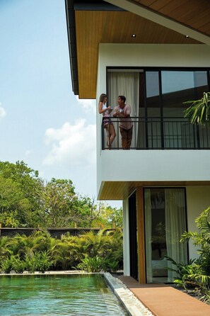 A modern two-story building with a balcony, a swimming pool, and lush greenery.