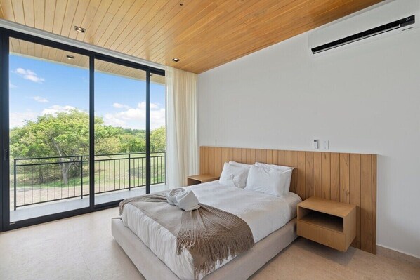 A modern bedroom with a large bed, wooden headboard, and a view of greenery through floor-to-ceiling windows.