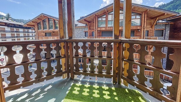 A wooden balcony with a patterned railing, a green artificial turf area, and buildings with wooden facades in the background. - Copalme - appartement - 5/7 pers - Centre (Les Gets)