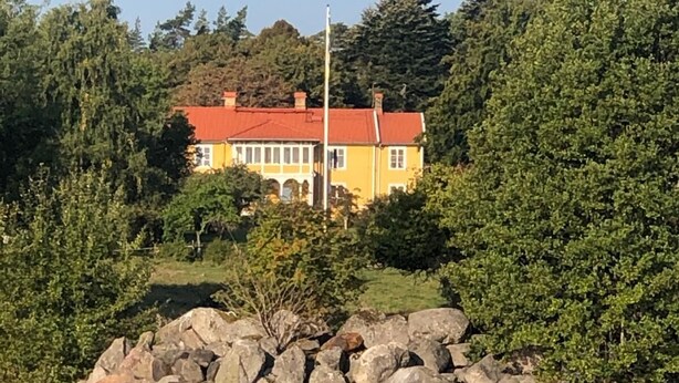 A yellow house with a red roof, surrounded by trees and rocks, reflecting in a calm body of water.