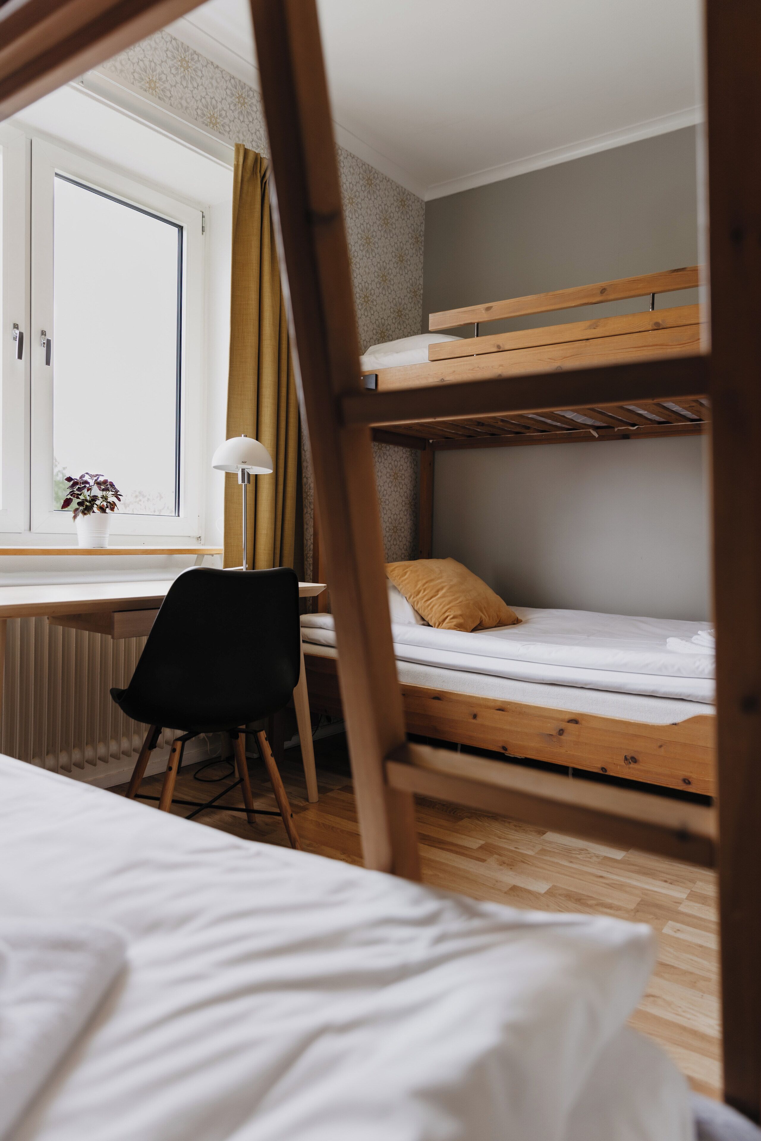 A bunk bed room with a desk and chair, a window with curtains, and a potted plant.