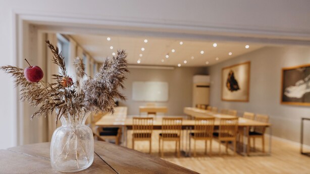 A dining area with a wooden table, chairs, and a clear glass vase with dried plants and a red fruit on the table. - Vassbo Logi & Event (Falun)