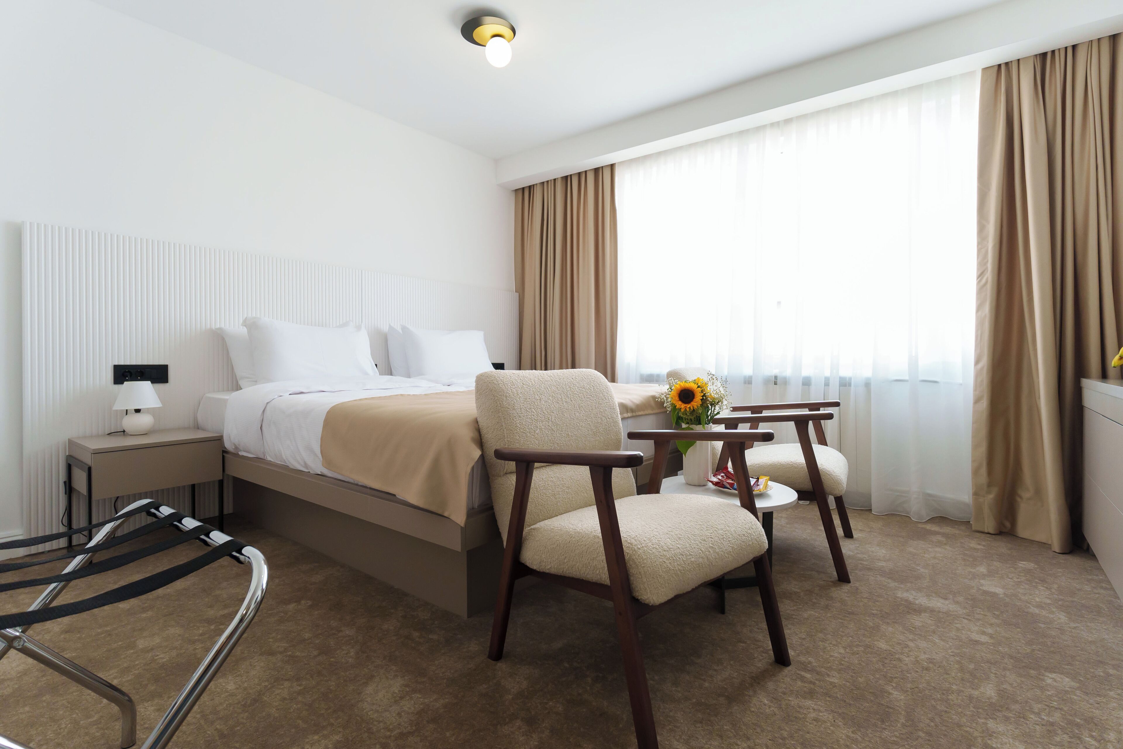 Une chambre d’hôtel avec un lit, une chaise, une petite table sur laquelle se trouve un vase, et une lampe.