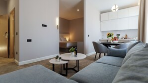 Un salon moderne avec un canapé gris, une table basse ronde et une petite table d’appoint agrémentée de plantes. On aperçoit un coin cuisine avec une table à manger et des chaises à travers une porte ouverte.