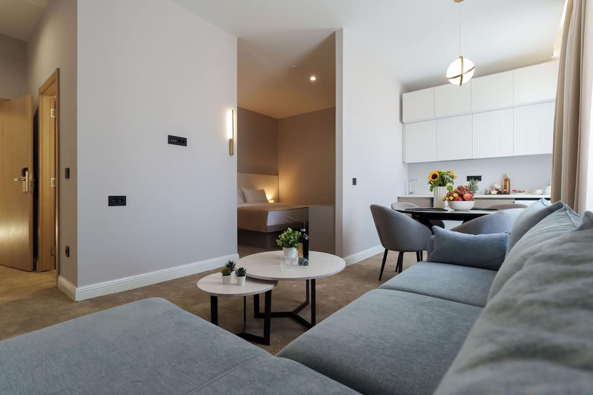 Ein modernes Wohnzimmer mit einem grauen Sofa, einem runden Couchtisch und einem kleinen Beistelltisch mit Pflanzen. Durch einen offenen Durchgang ist eine Küchenzeile mit Esstisch und Stühlen zu sehen.