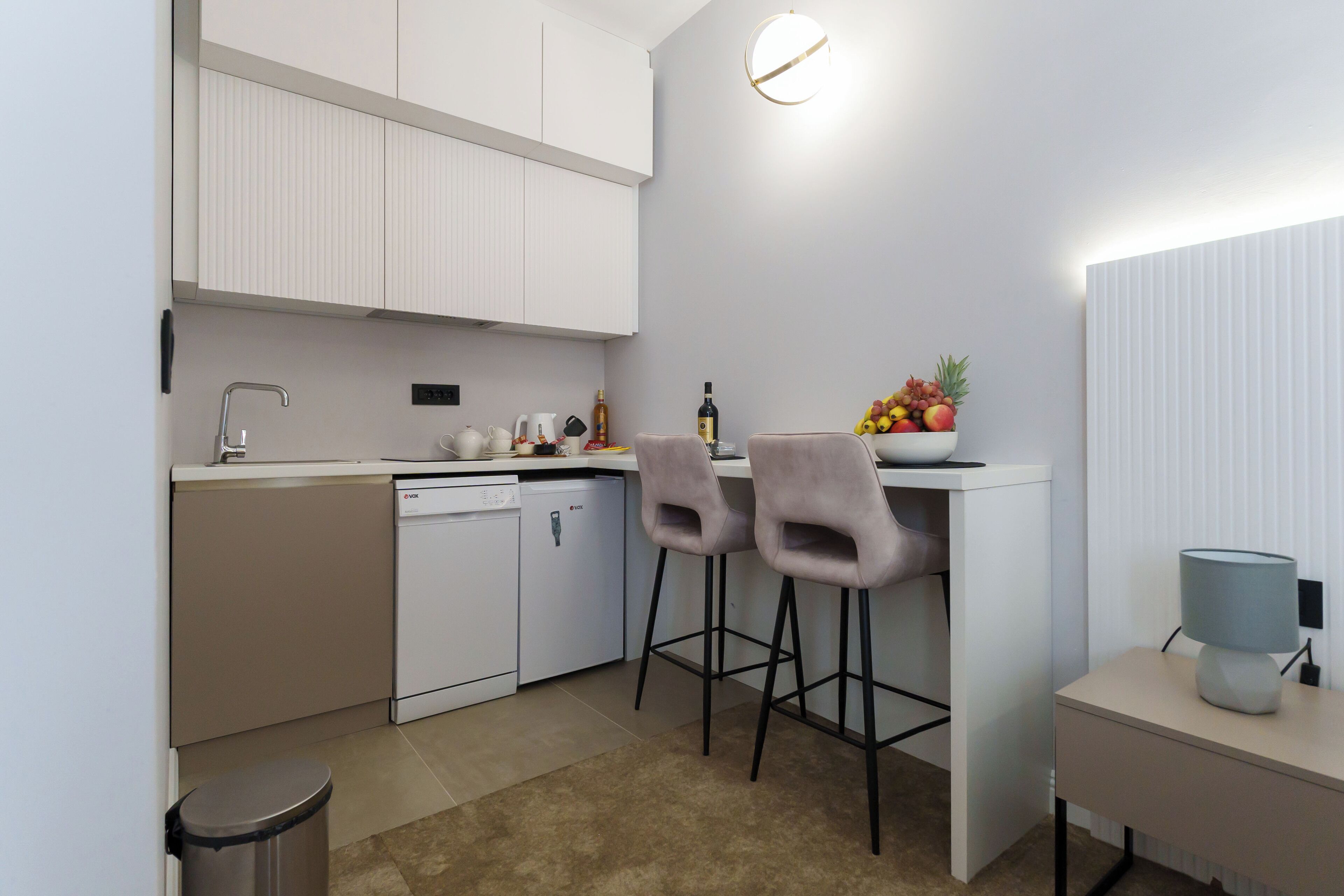 Una cocina compacta con muebles blancos, un lavavajillas, un fregadero y dos taburetes de bar.