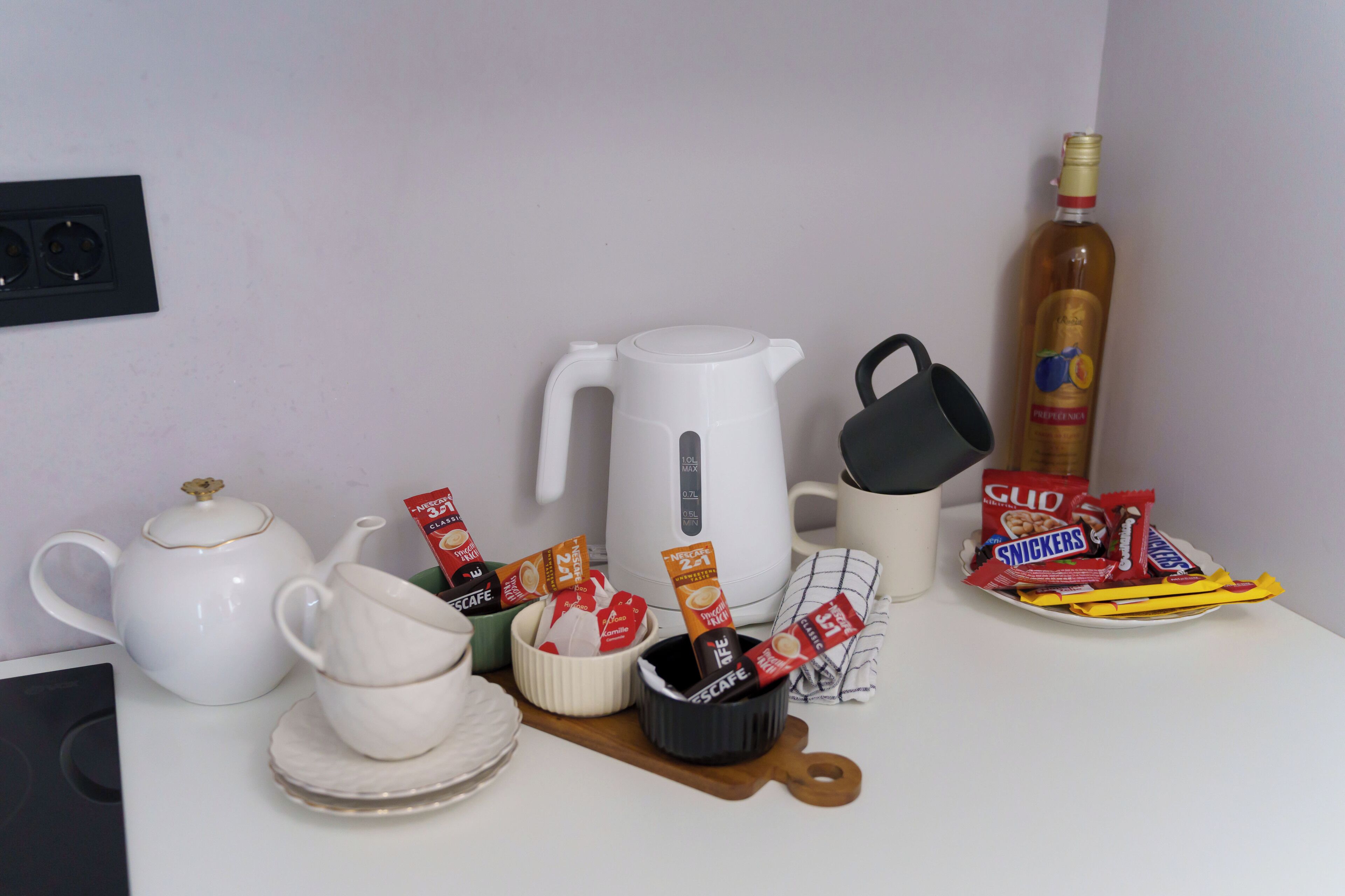 Un bancone cucina con un bollitore bianco, una macchina per il caffè, una bottiglia d'olio d'oliva e diverse confezioni di snack.