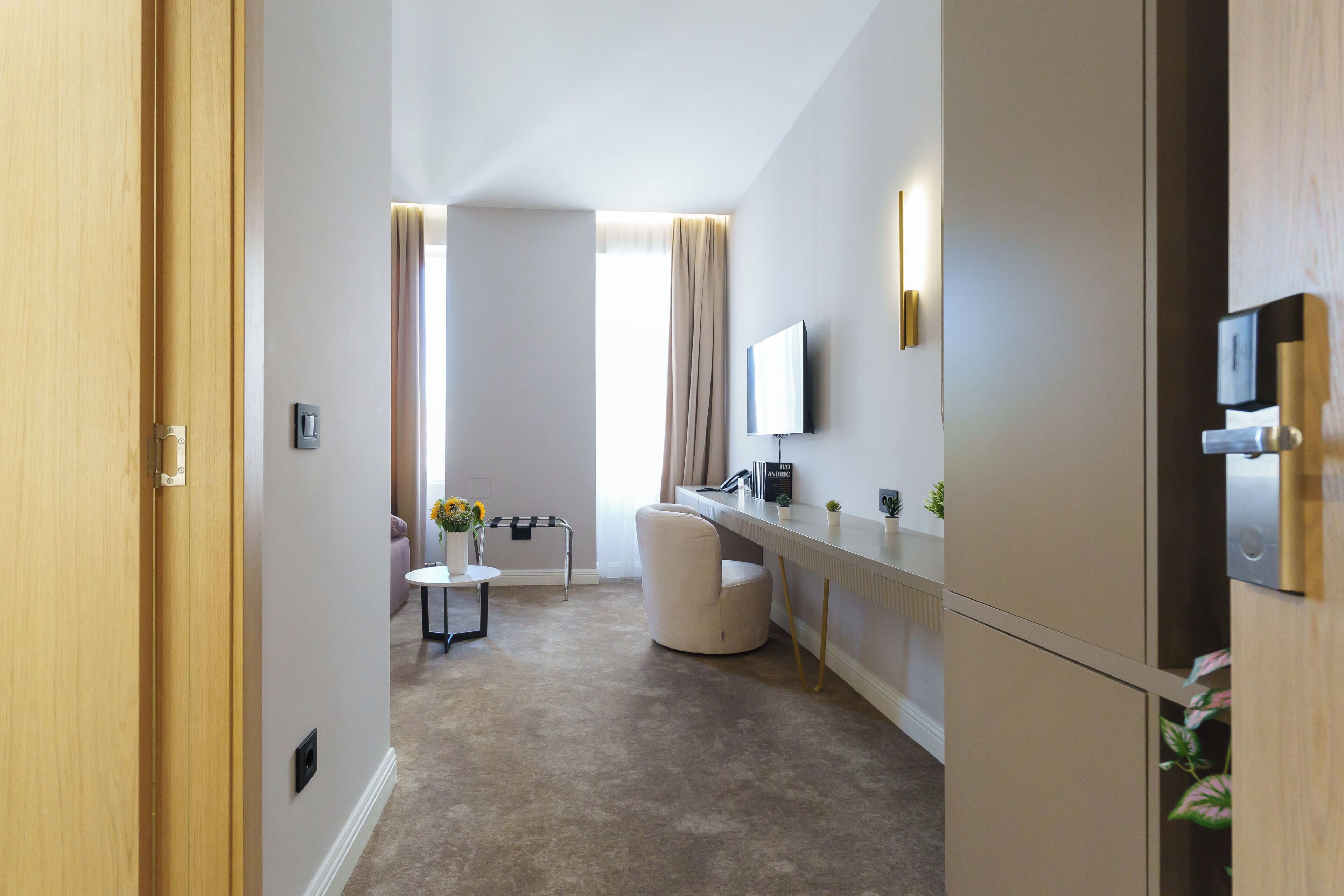 Une chambre d’hôtel moderne avec une porte en bois, un téléviseur à écran plat fixé au mur, une petite table sur laquelle est posé un vase de fleurs, une chaise et une grande fenêtre avec des rideaux.