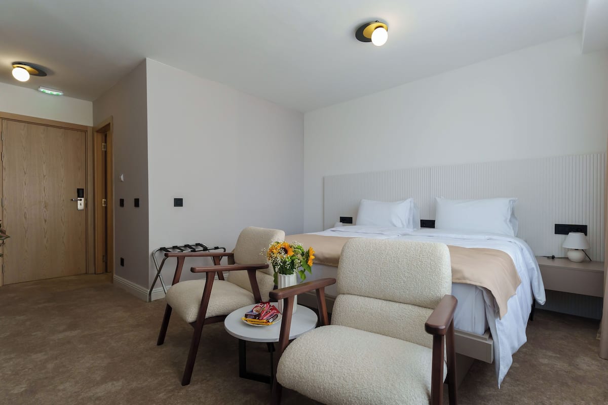 Ein Hotelzimmer mit einem Bett, zwei Sesseln, einem kleinen Tisch mit Blumenvase und einem Nachttisch mit Lampe.