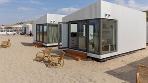 A modern beachside building with large glass windows, a wooden deck, and wicker furniture. - Beach House in Zandvoort With Sea View (Zandvoort)