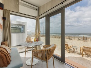 A modern room with a large glass door leading to a beach view, featuring a round table, wicker chairs, and a sofa with a throw blanket.