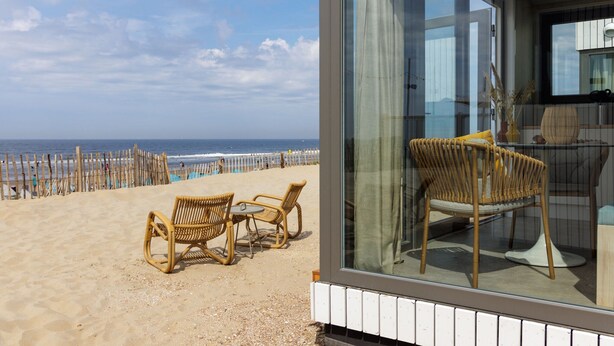 A beachside view with wicker chairs and a table, a glass wall, and a view of the ocean. - Beach House in Zandvoort With Sea View (Zandvoort)