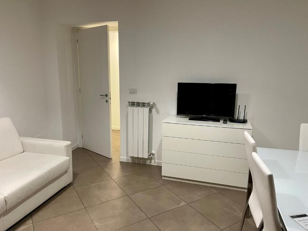A room with a white sofa, a TV on a white cabinet, a dining table with chairs, and a door leading to another room.
