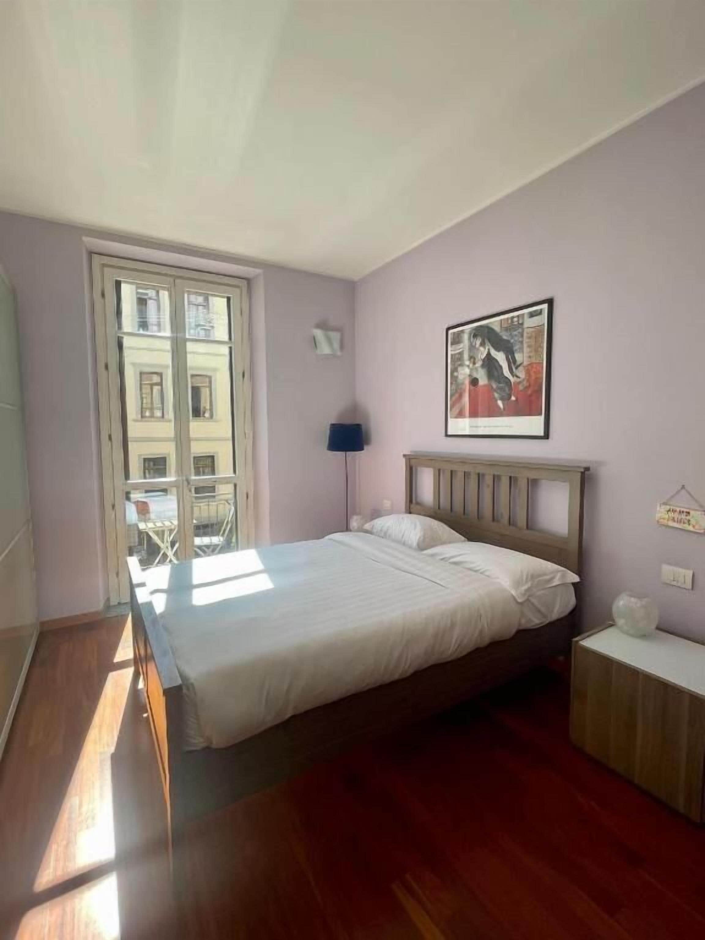 Une chambre à coucher avec un lit, une table de chevet, une lampe, un tableau encadré et une vue sur un immeuble par la fenêtre.