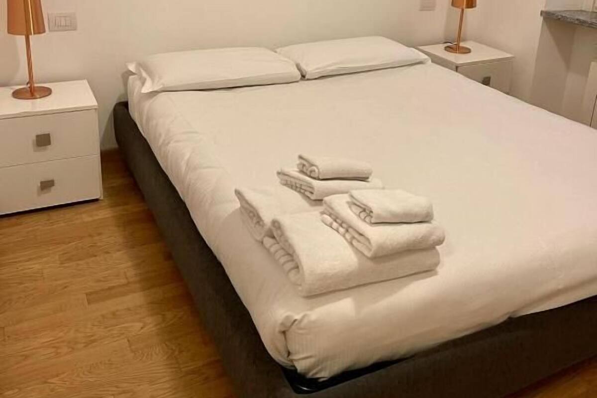 A neatly made bed with white linens and folded towels, two bedside lamps, and a wooden floor.