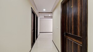 A hallway with a wooden door on the right, a white door on the left, and a white wall with a light switch. - Hotel Shashwat (Gorakhpur)