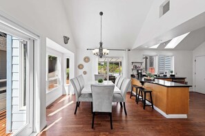 Una cocina amplia con una isla central, un comedor con candelabro y vistas a un jardín a través de ventanas grandes.