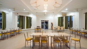 A spacious dining area with multiple tables and chairs, ceiling fans, and large windows with curtains. - Corbett Holiday Forest Resort (Belparao)