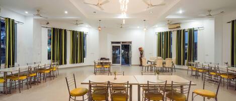A spacious dining area with multiple tables and chairs, ceiling fans, and large windows with curtains.