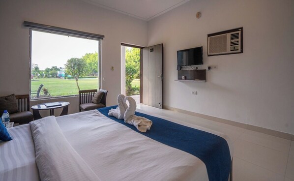 A hotel room with a large bed, a TV mounted on the wall, a chair, a small table, and a view of a green landscape through the window. - Corbett Holiday Forest Resort (Belparao)