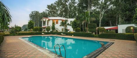 A swimming pool with a tiled deck, surrounded by a well-maintained garden and a large building with a red roof.