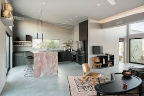 Contemporary living space with a minimalist design, featuring a kitchen with dark cabinetry and a stainless steel refrigerator, a dining area with a table and chairs, and a large window with an outdoor view.