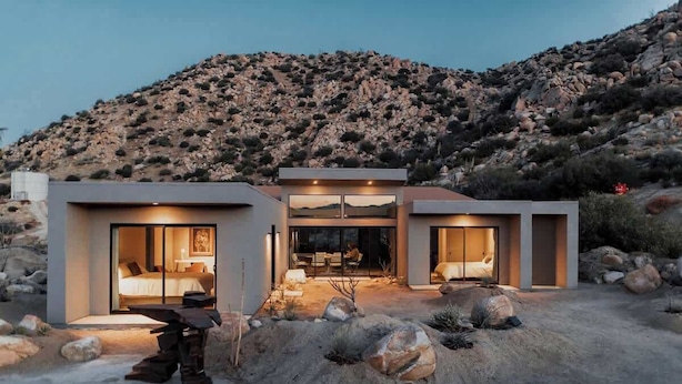 A modern, two-story house with large glass windows and doors, a patio area, and a mountainous backdrop.