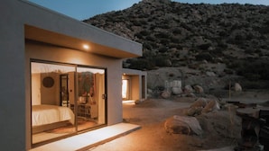 A modern, well-lit building with a glass door leading to a bedroom. The exterior features a minimalist design with large rocks and a desert landscape in the background.