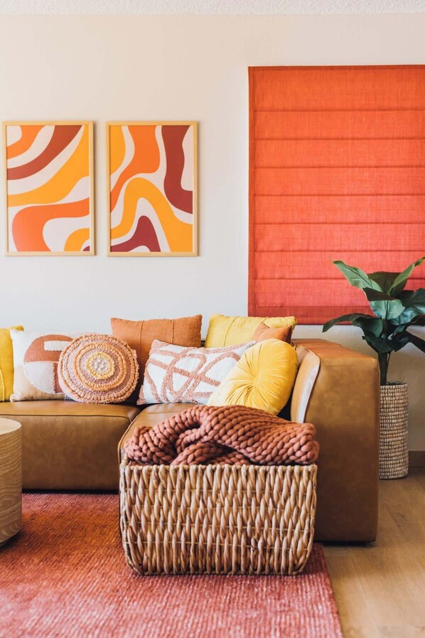 Una sala de estar acogedora con un sofá marrón, cojines coloridos, un reposapiés tejido y dos cuadros abstractos en la pared.