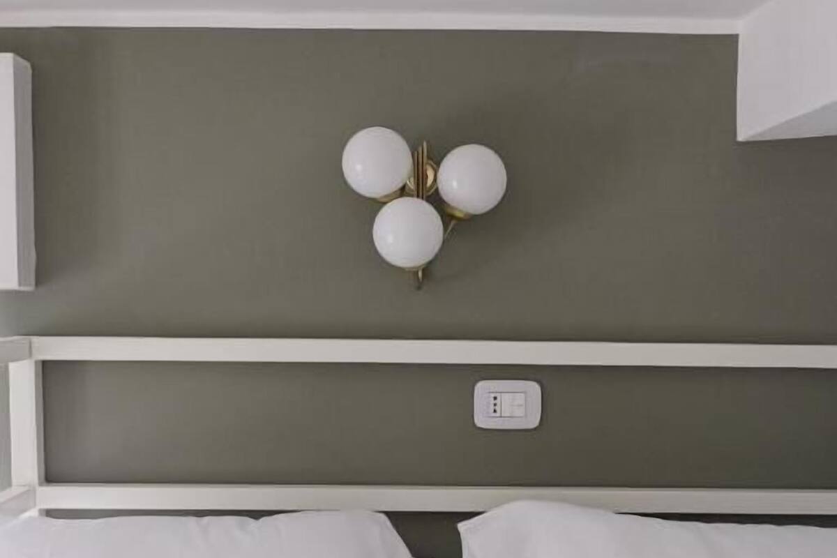 A bed with white linens, a white headboard, and a wall-mounted light fixture with three spherical bulbs.
