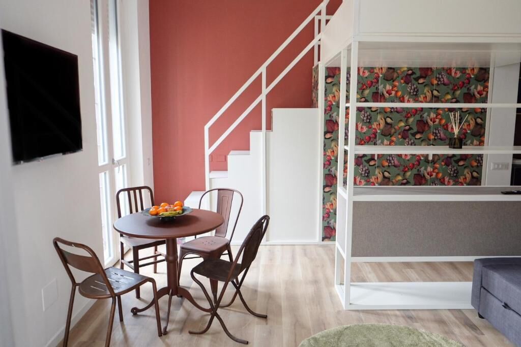 Un comedor con una mesa redonda y cuatro sillas, una escalera con barandillas blancas y una estantería con papel tapiz floral.