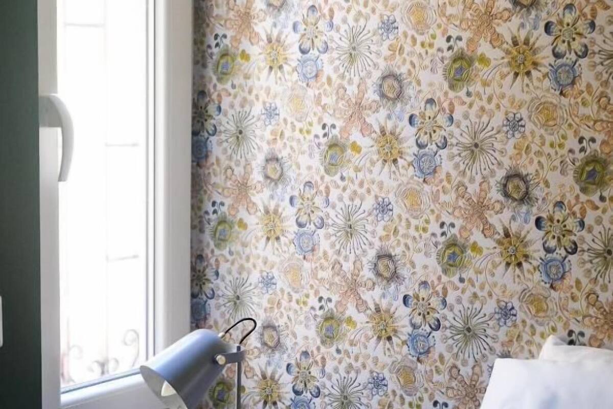A bedroom with a bed, a bedside lamp, a window, and a wall with floral wallpaper.