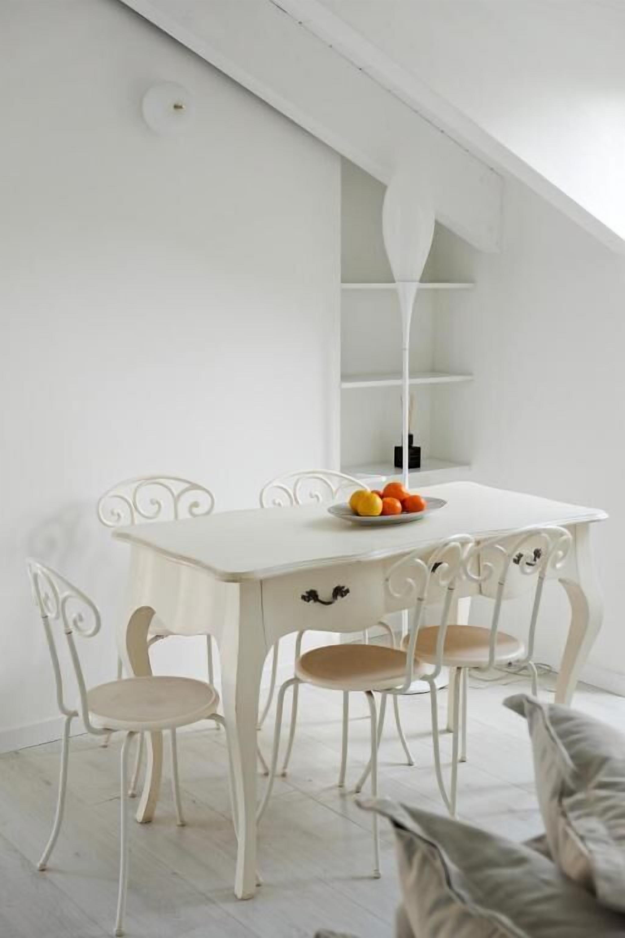 Een witte eettafel met vier stoelen, een witte hanglamp en een klein plankje met sinaasappels.