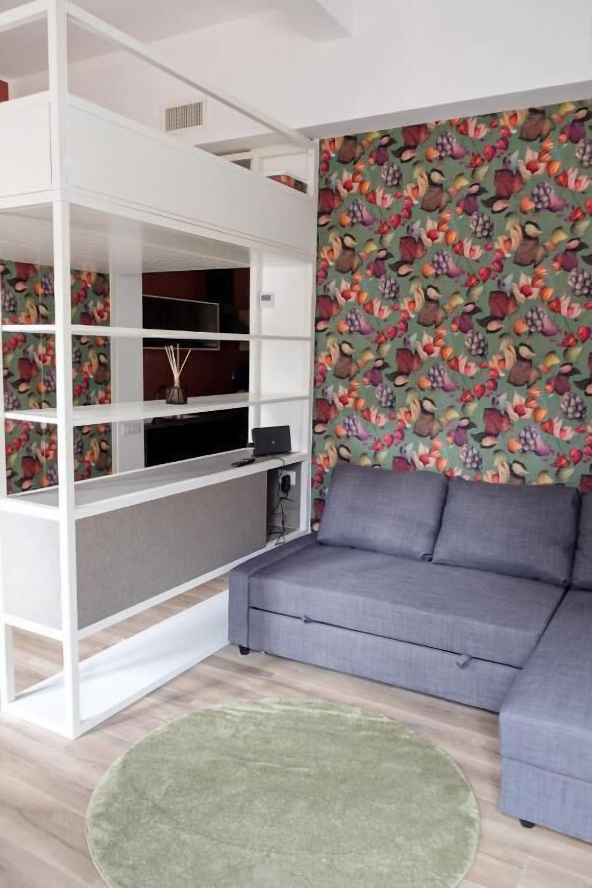Ein modernes Wohnzimmer mit einer grauen Eckcouch, einer weißen Regaleinheit und ornamentalem Wandpapier.
