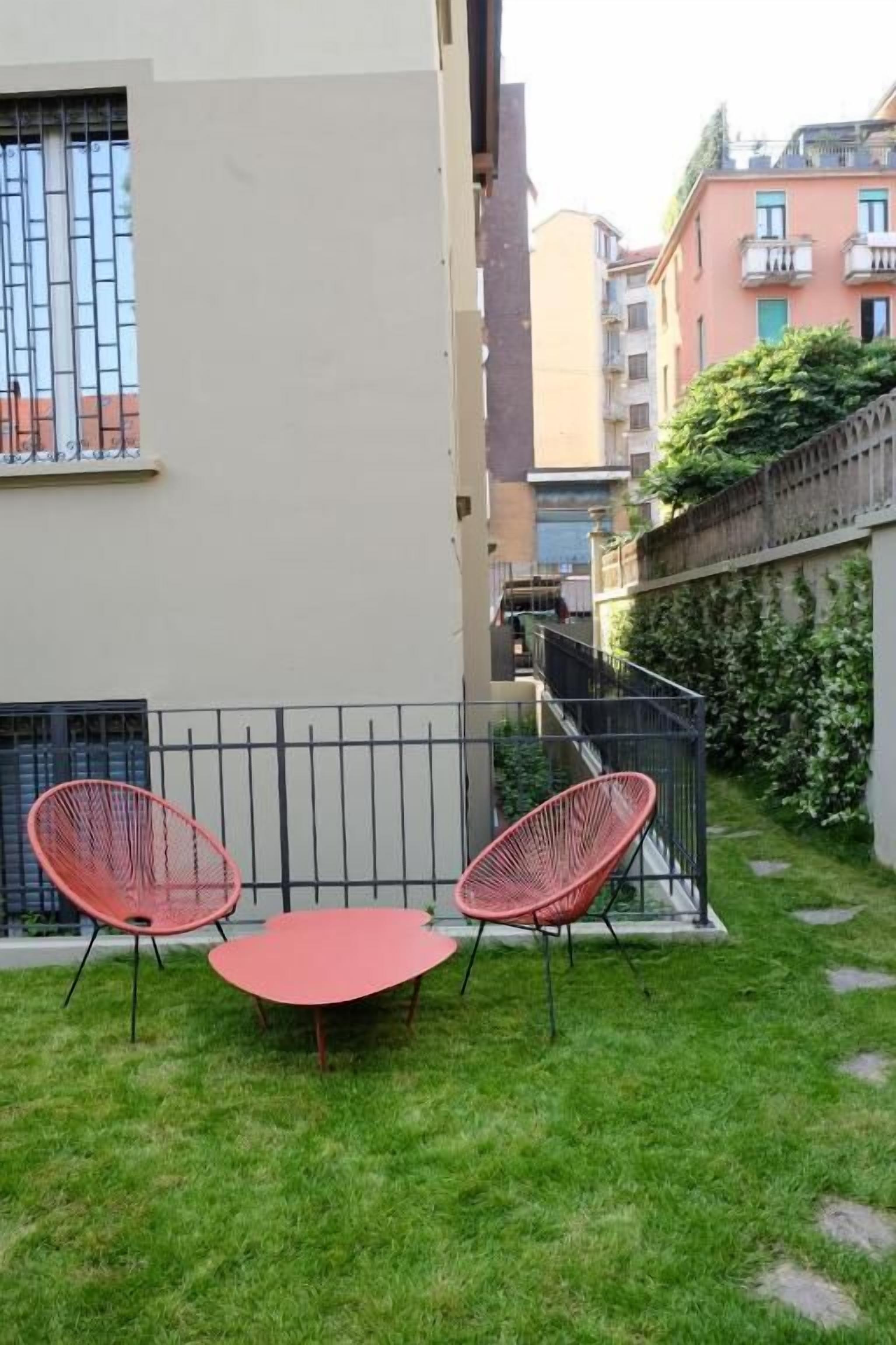 Un pequeño patio al aire libre con una mesa redonda y dos sillas rojas, rodeado por una cerca de metal negro bajo y una zona con césped.
