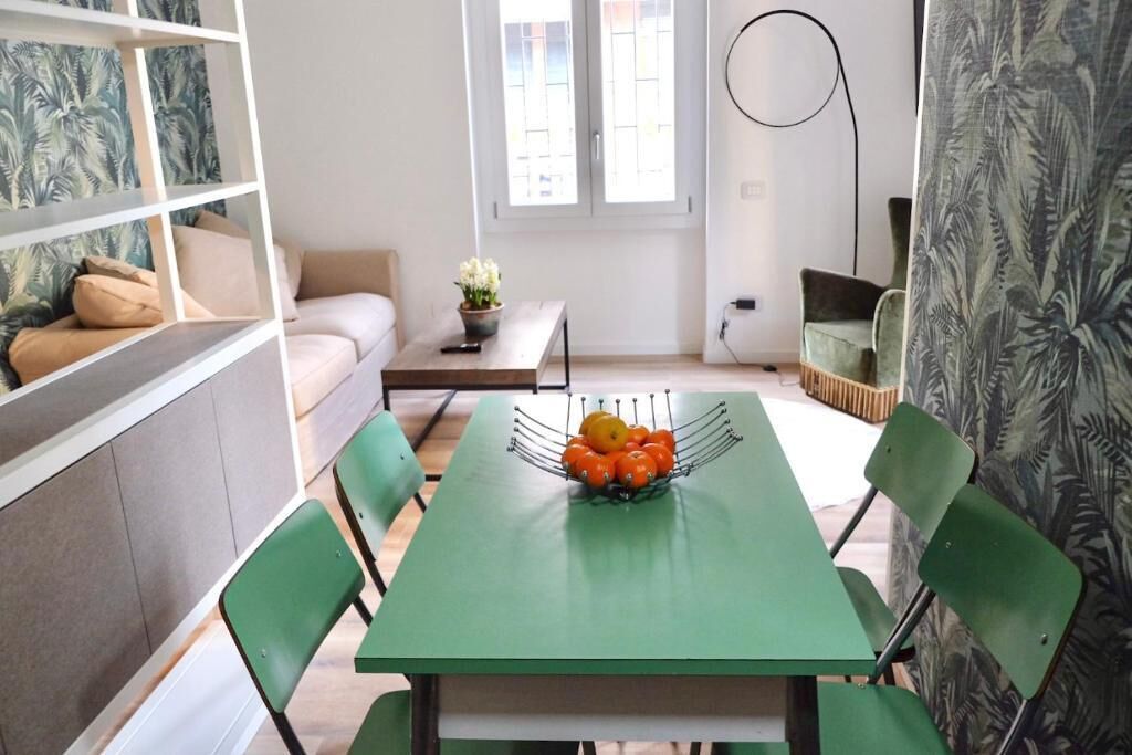 A dining area with a green table, chairs, and a fruit bowl. There is a sofa and a window with blinds in the background.