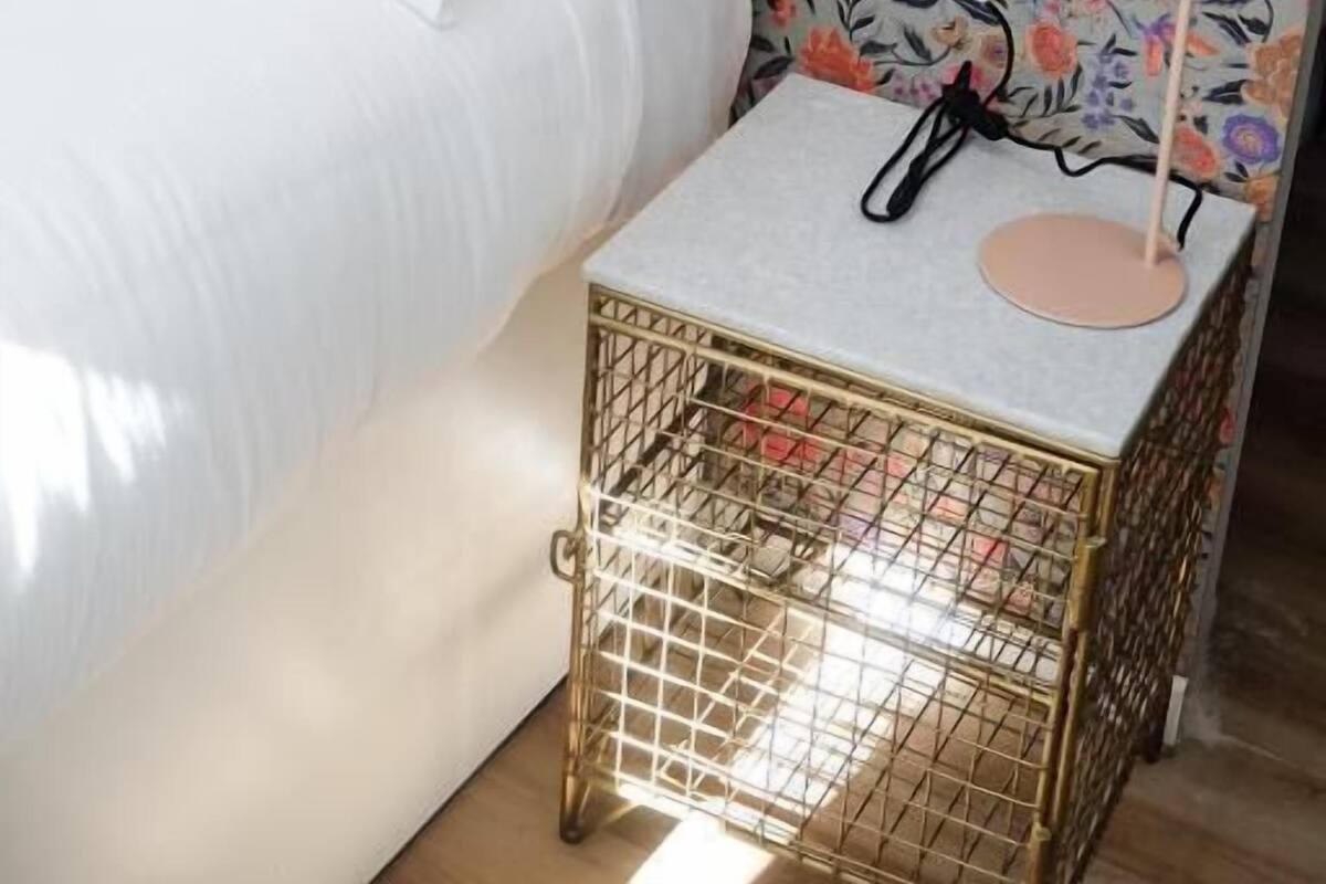 A neatly made bed with white linens, a floral wallpaper headboard, and a bedside table with a lamp and a small gold-framed basket.