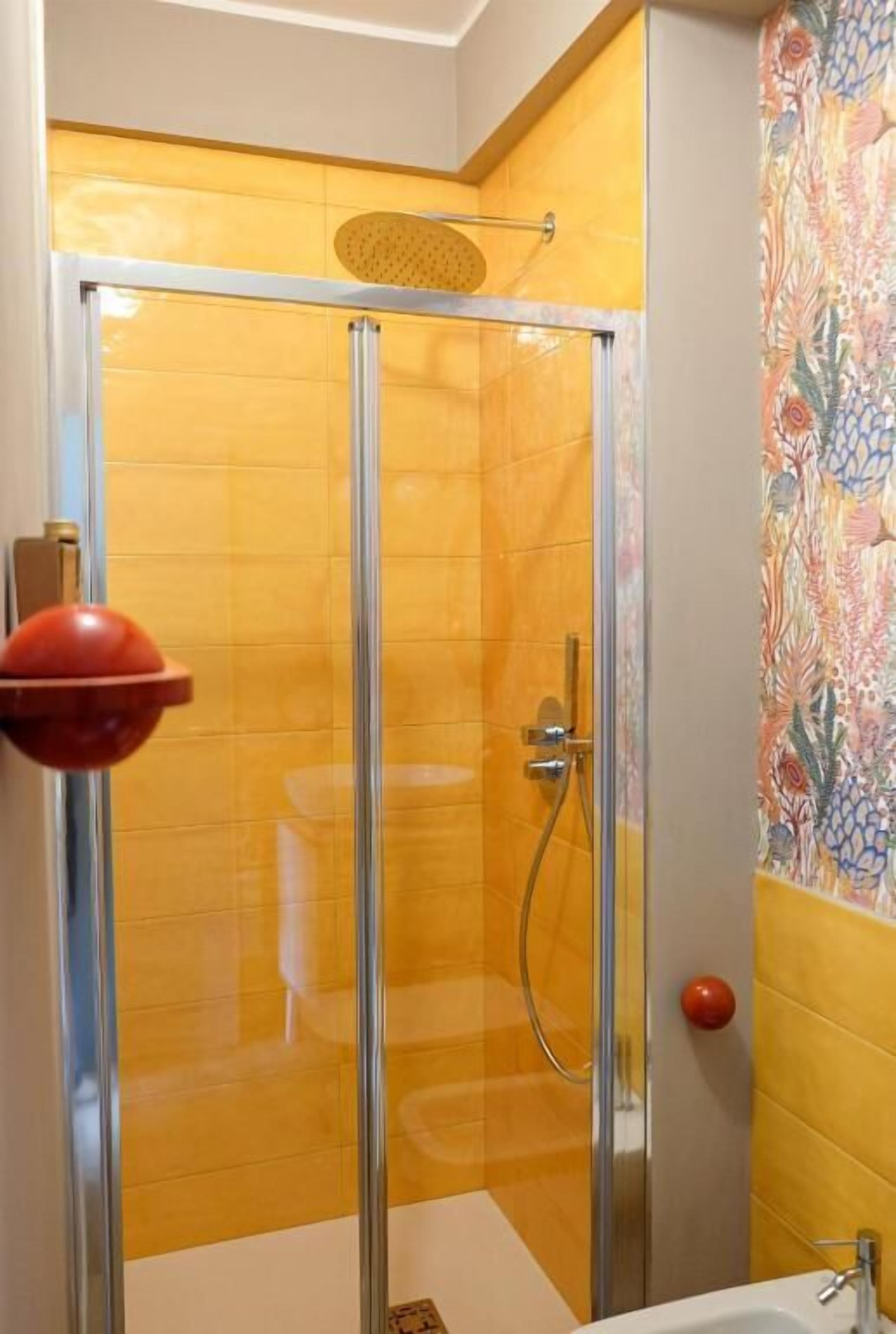 Une douche fermée par des parois de verre, dotée d’un pommeau de douche à main, d’un porte-savon rouge et d’un mur décoratif recouvert de carreaux.