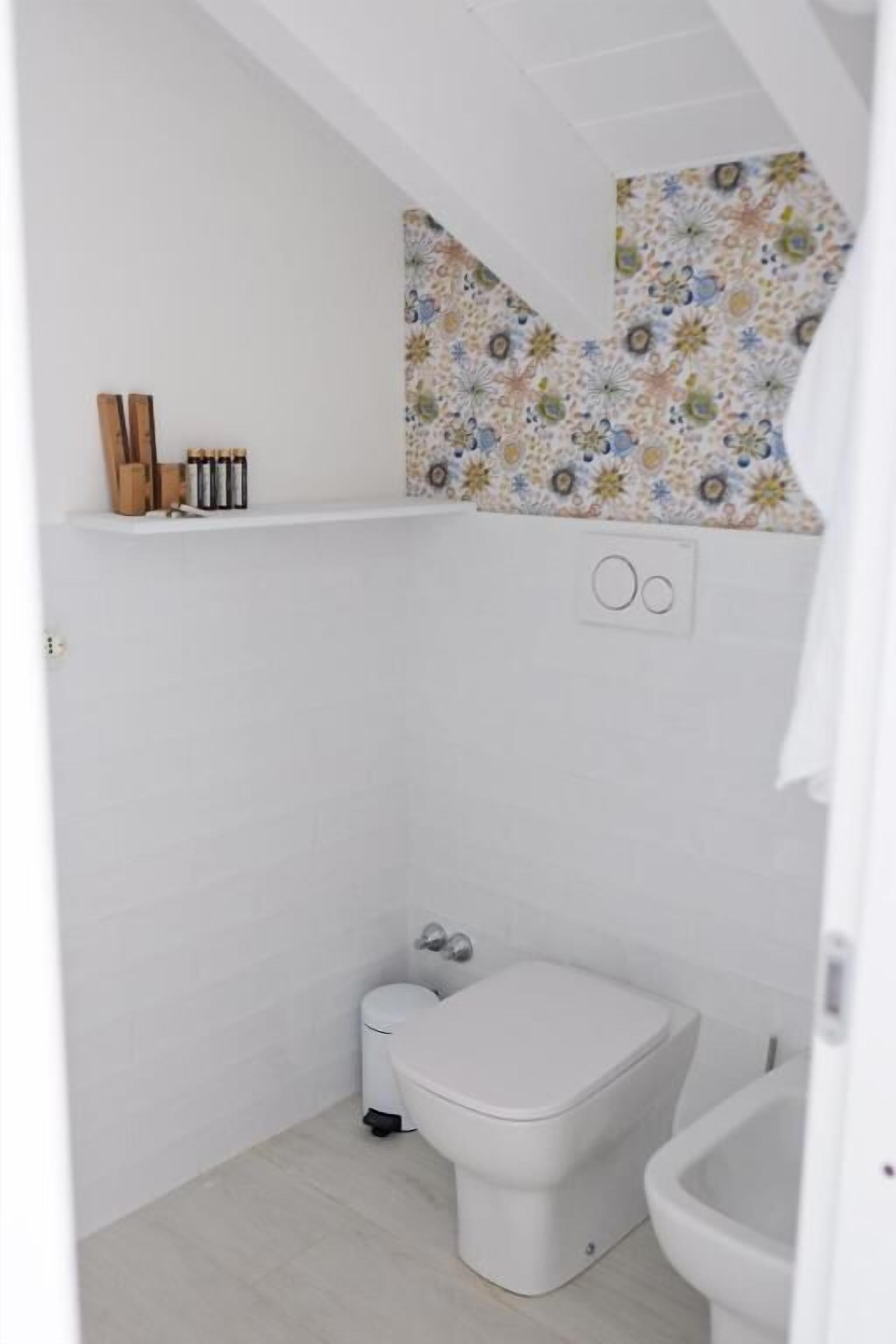 A small, white bathroom with a toilet, bidet, and a shelf with toiletries.