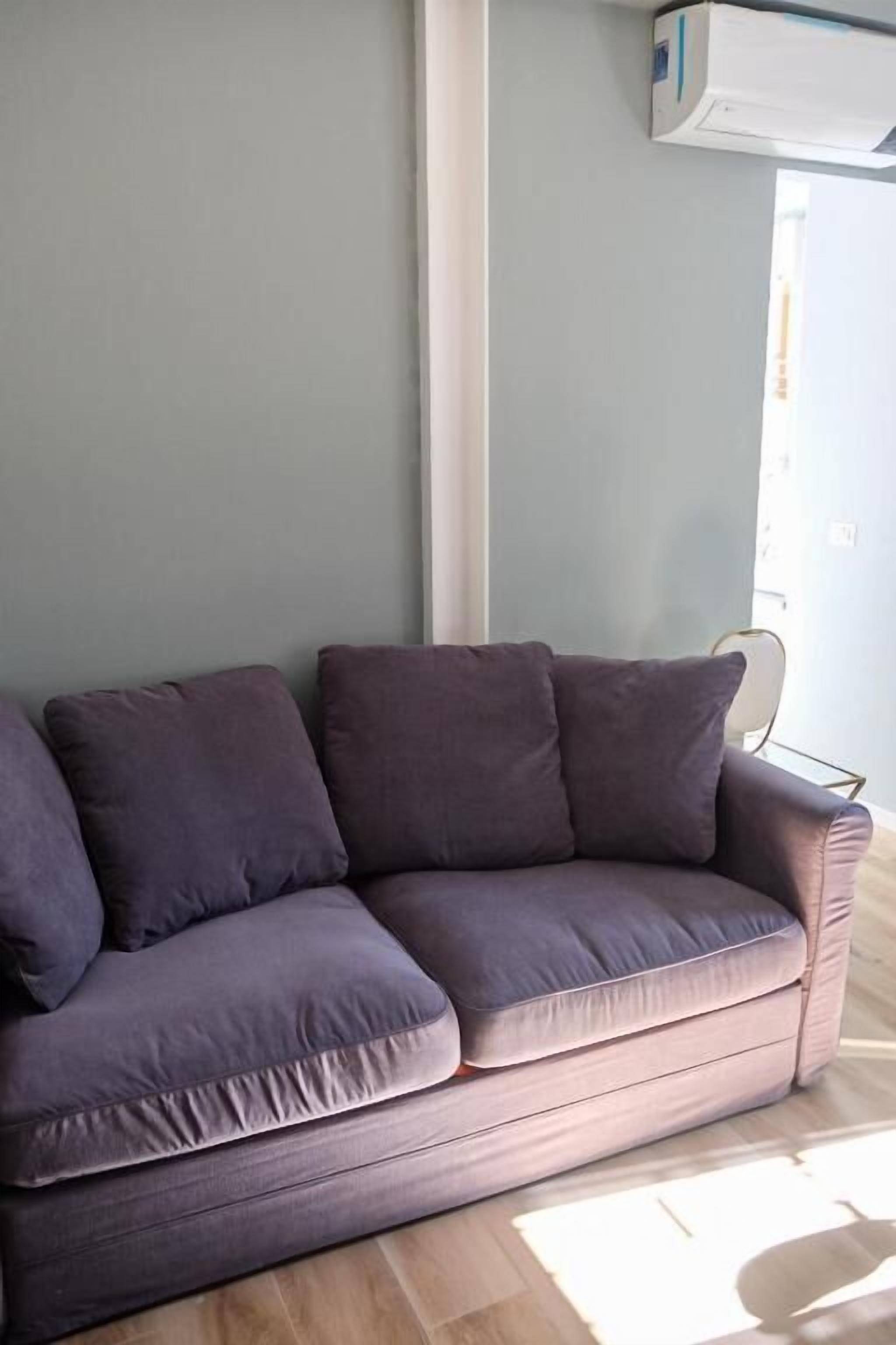 A grey sofa with cushions in a room with light grey walls and a window.