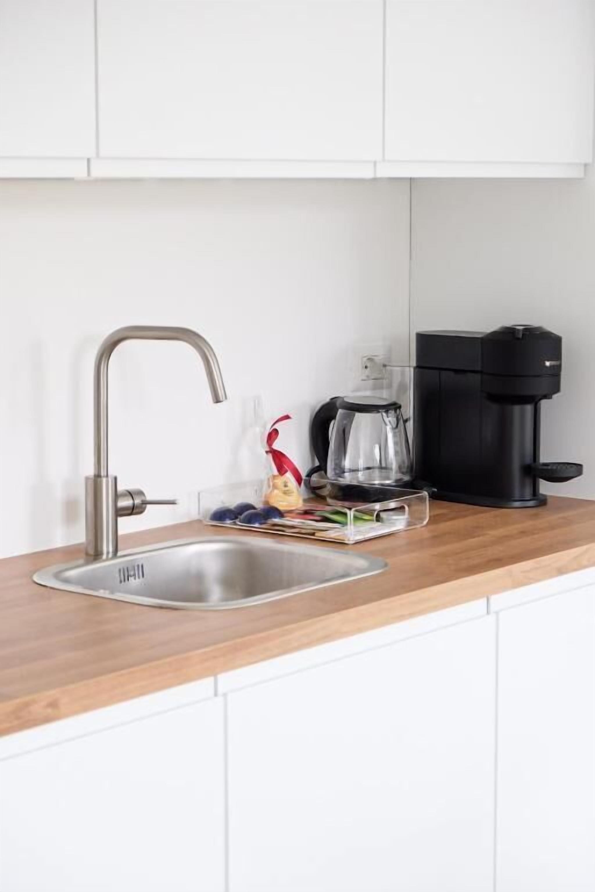 Una cocina con un lavamanos de acero inoxidable, una cafetera y una jarra de cristal transparente sobre la encimera.