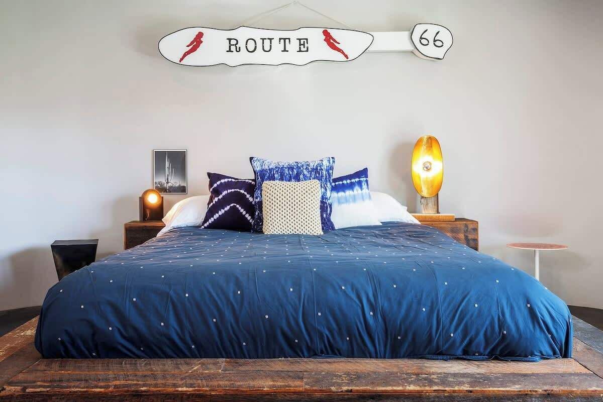 A bedroom with a large bed, a nightstand with a lamp, a wall-mounted sign reading "ROUTE 66", and a framed picture on the wall.