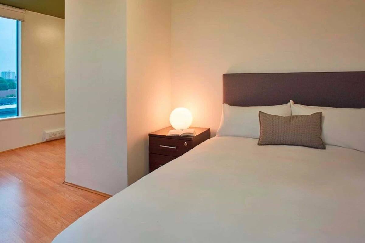 A hotel room with a large bed, a bedside table with a lamp, and a view of the city through a window.