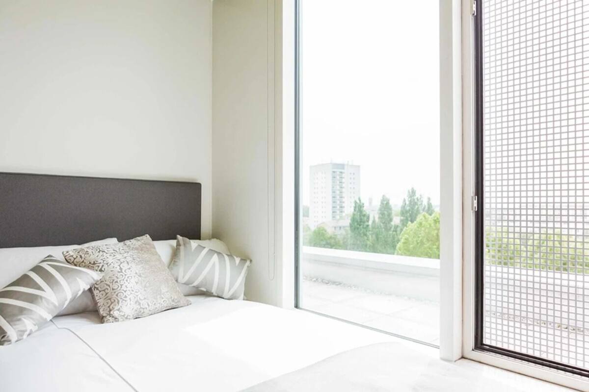 A modern bedroom with a large bed, a grey headboard, and a view of a cityscape through a sliding glass door.