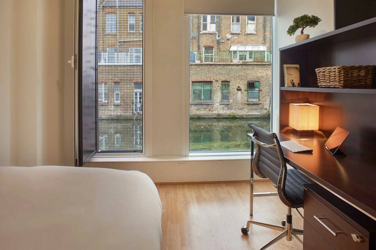 A hotel room with a bed, a desk with a chair, a lamp, a small plant, and a view of a canal and buildings.