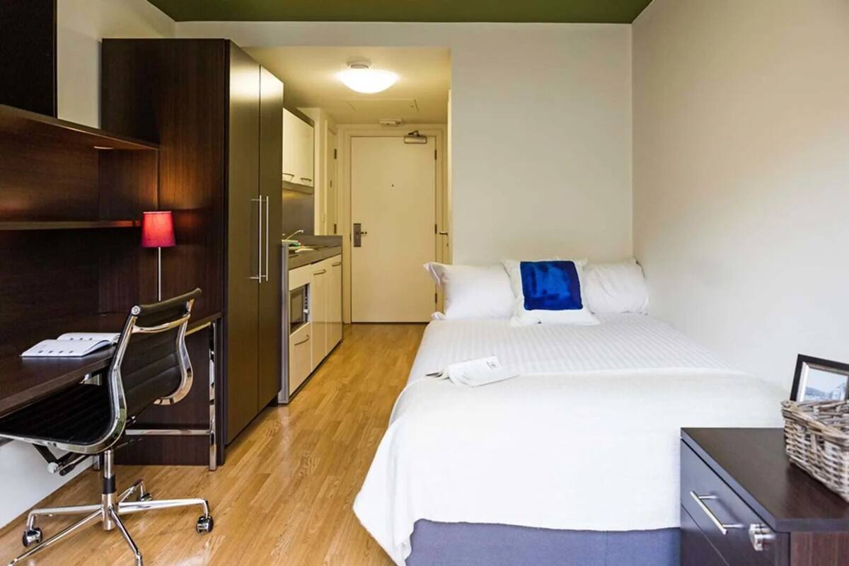 A hotel room with a bed, a desk with a chair, a wardrobe, and a kitchenette.