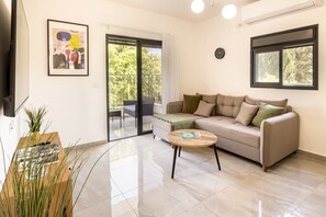 A living room with a sofa, a coffee table, a framed artwork, a sliding glass door, and a window. - MAMAD - Bahai Gardens Vista Apts by Sea N' Rent (Haifa)